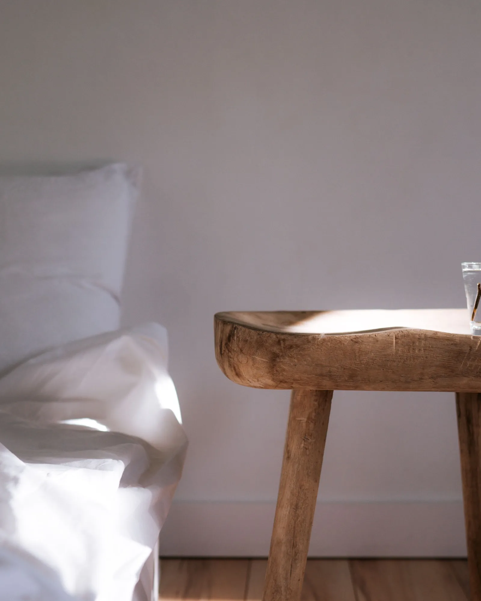 Chambre baignée de lumière au réveil : lit au linge blanc, tabouret de chevet en bois et verre d’eau